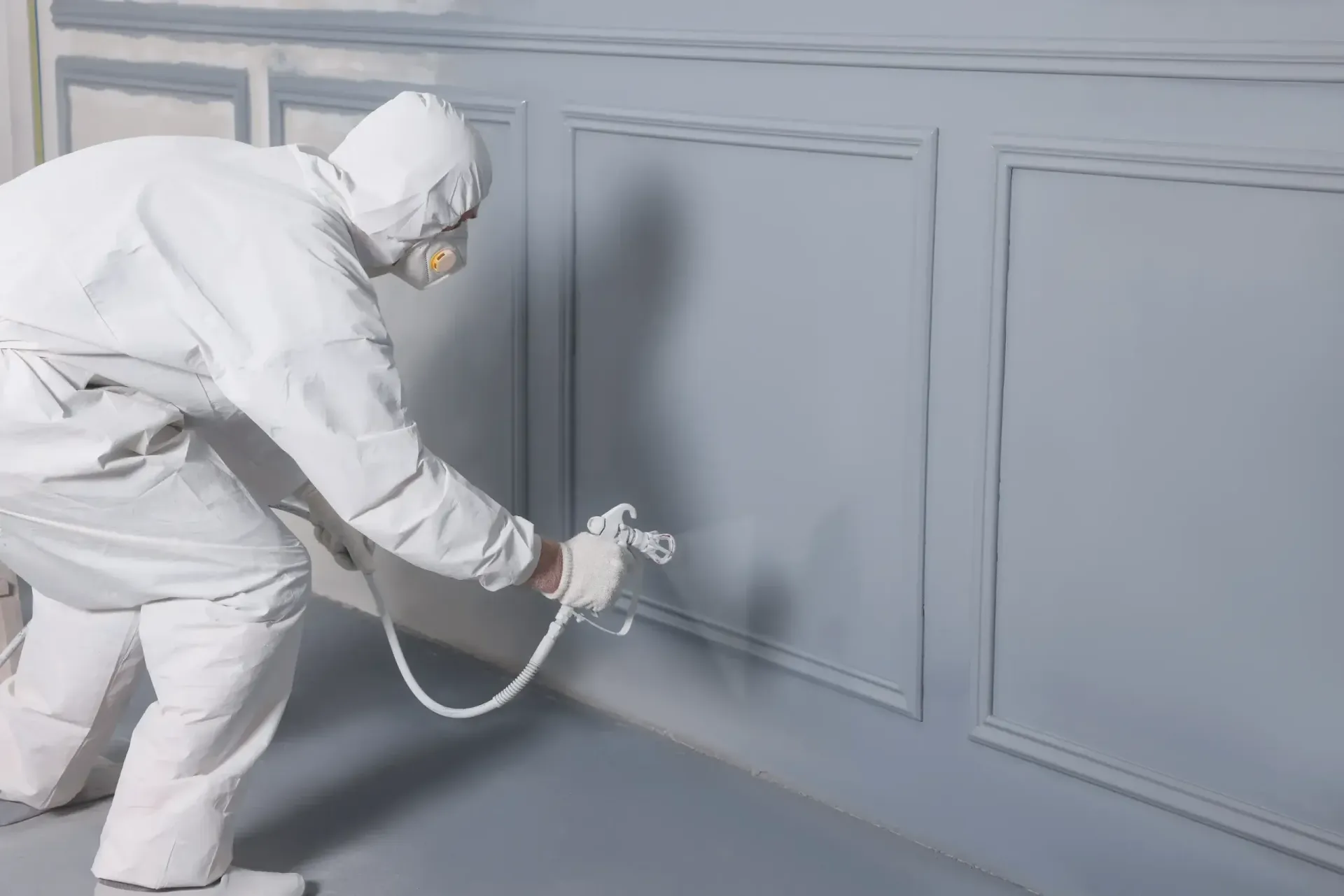 Person in protective suit spray painting gray paneling on a wall.