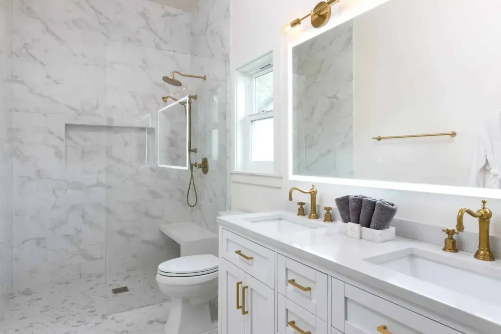 Bright white bathroom with gold fixtures, marble walls, double vanity, and walk-in shower.