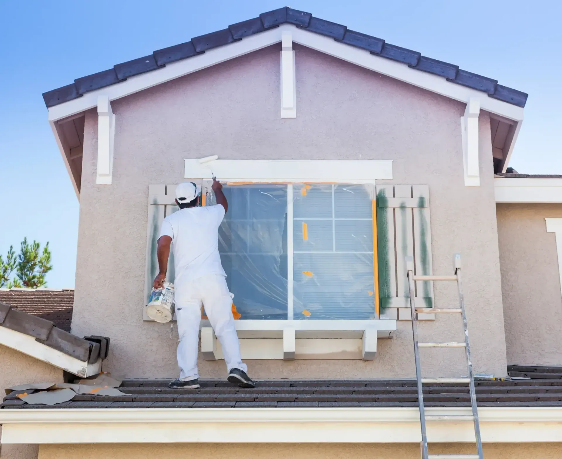 Painter in white overalls on a ladder painting window trim on a beige house. Blue sky.