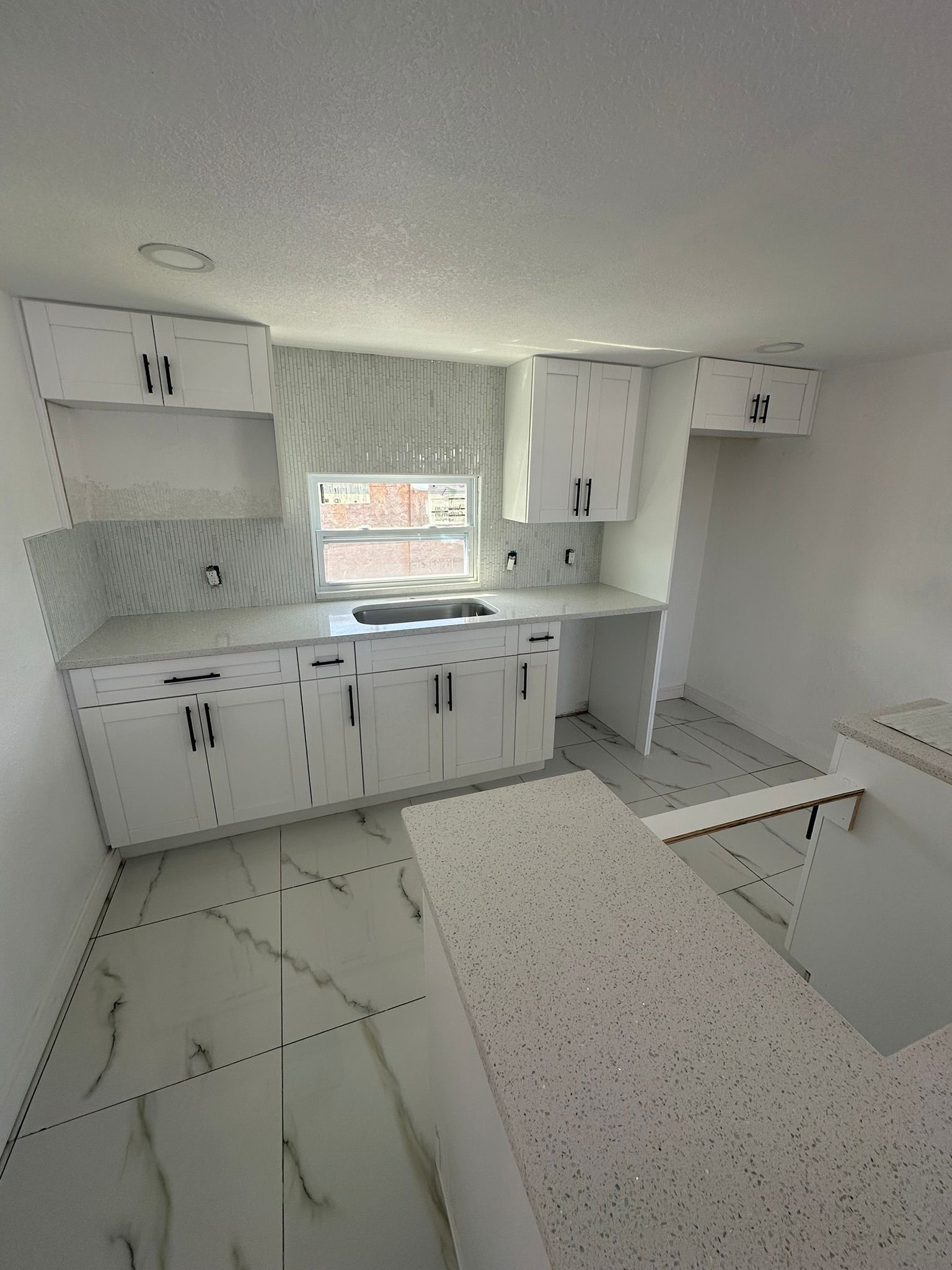 White kitchen with cabinets, countertops, and tile backsplash, with an island and window.