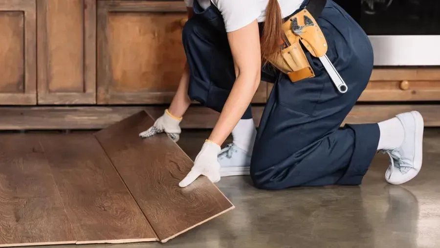 Person kneeling, installing wood flooring. Wearing gloves and work clothing in a kitchen setting.