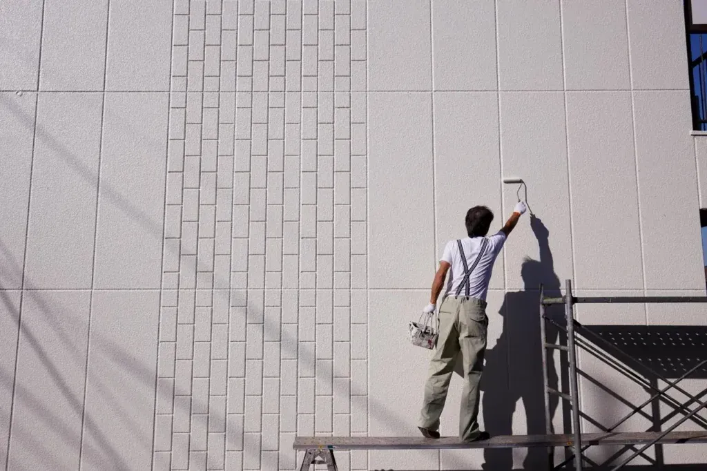 Painter on scaffolding, applying paint to exterior wall with a brick-like pattern. Bright sunlight, white and gray colors.