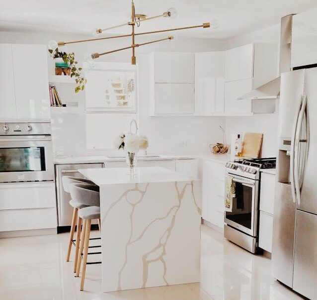 Modern white kitchen with quartz island, stainless steel appliances, and gold chandelier.
