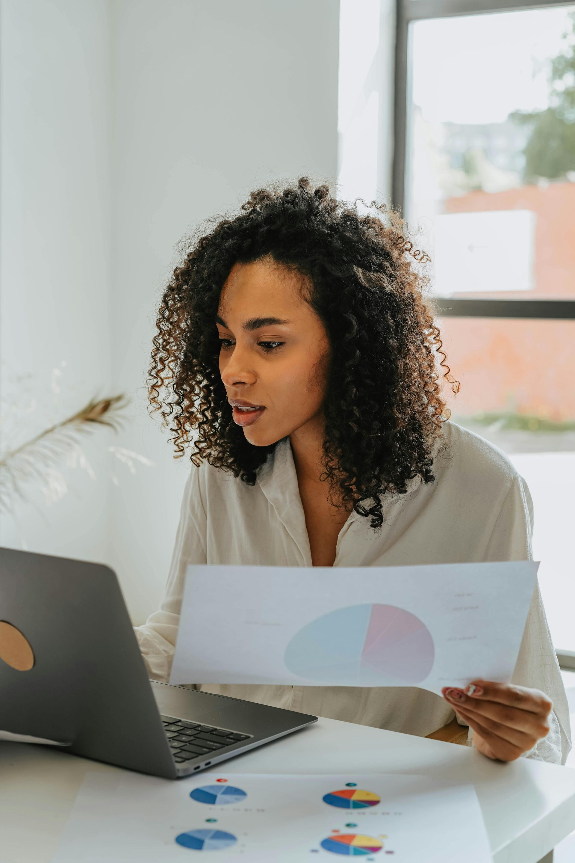 A woman is sitting at a desk looking at a laptop and holding a piece of paper.