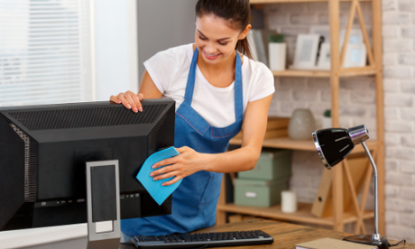 Person cleaning a desktop computer monitor with a blue cloth in a home office