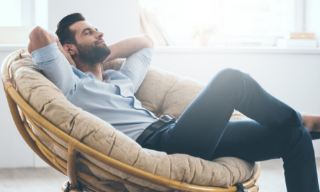 Man reclining in a cushioned chair by a bright window, resting with hands behind his head.