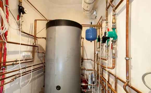 A large, grey cylindrical water storage tank centered in a mechanical room, surrounded by copper pipes and plumbing pumps.