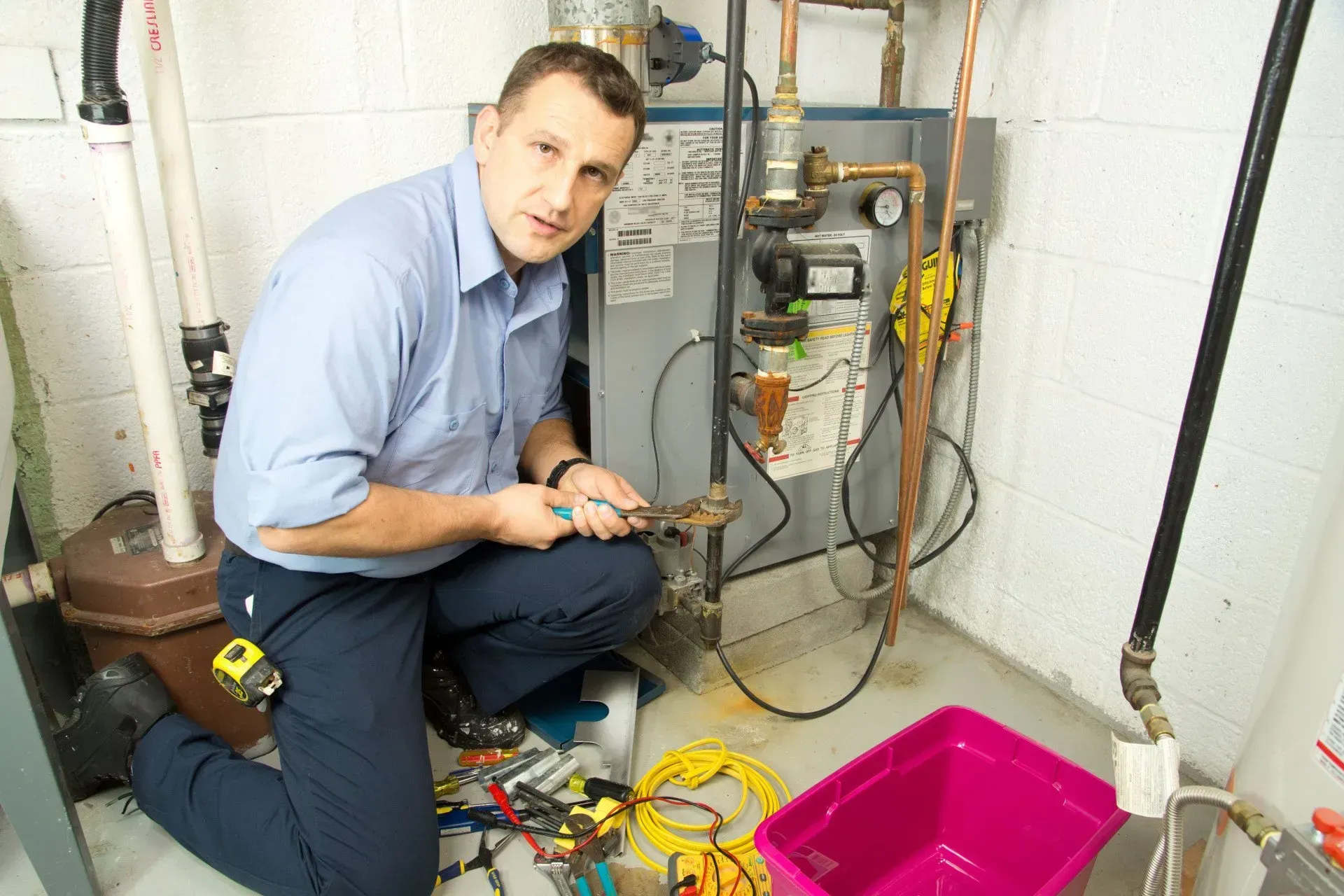 A professional worker repairs a boiler in a utility room while kneeling beside a pink bin and various tools.