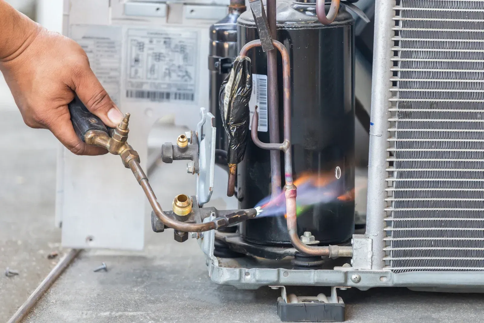 A technician uses a blowtorch to solder a metal pipe on an outdoor air conditioning compressor unit.