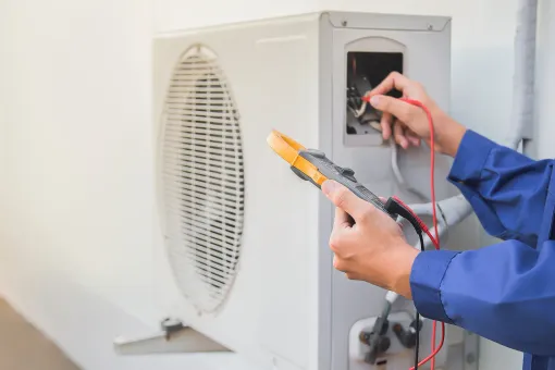 Technician in a blue uniform testing an outdoor AC unit’s wiring using a digital multimeter.
