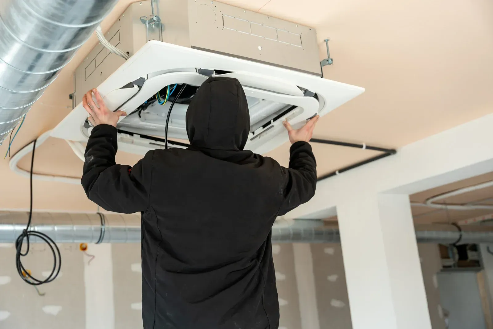 A person wearing a black hoodie installing or repairing a white ceiling-mounted HVAC unit in an unfinished room.