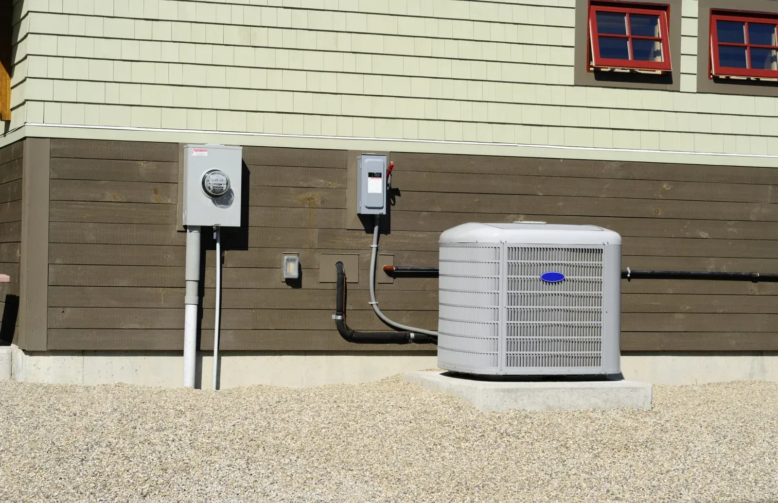 An outdoor AC unit sits on a gravel patch against a building wall with electrical equipment boxes nearby.