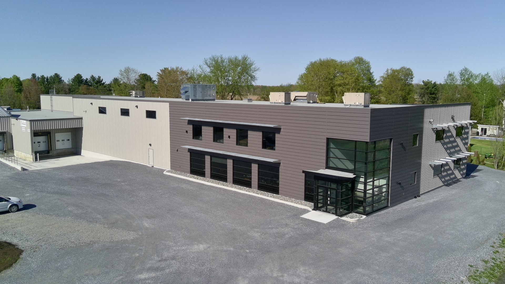 Bâtiment moderne avec parking en gravier gris, ciel bleu et arbres en arrière-plan.