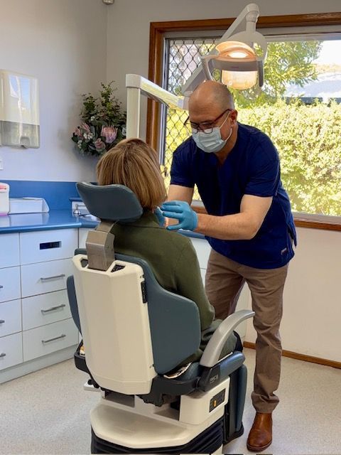 Dentist wearing a mask and gloves treats a seated patient in a bright dental office.