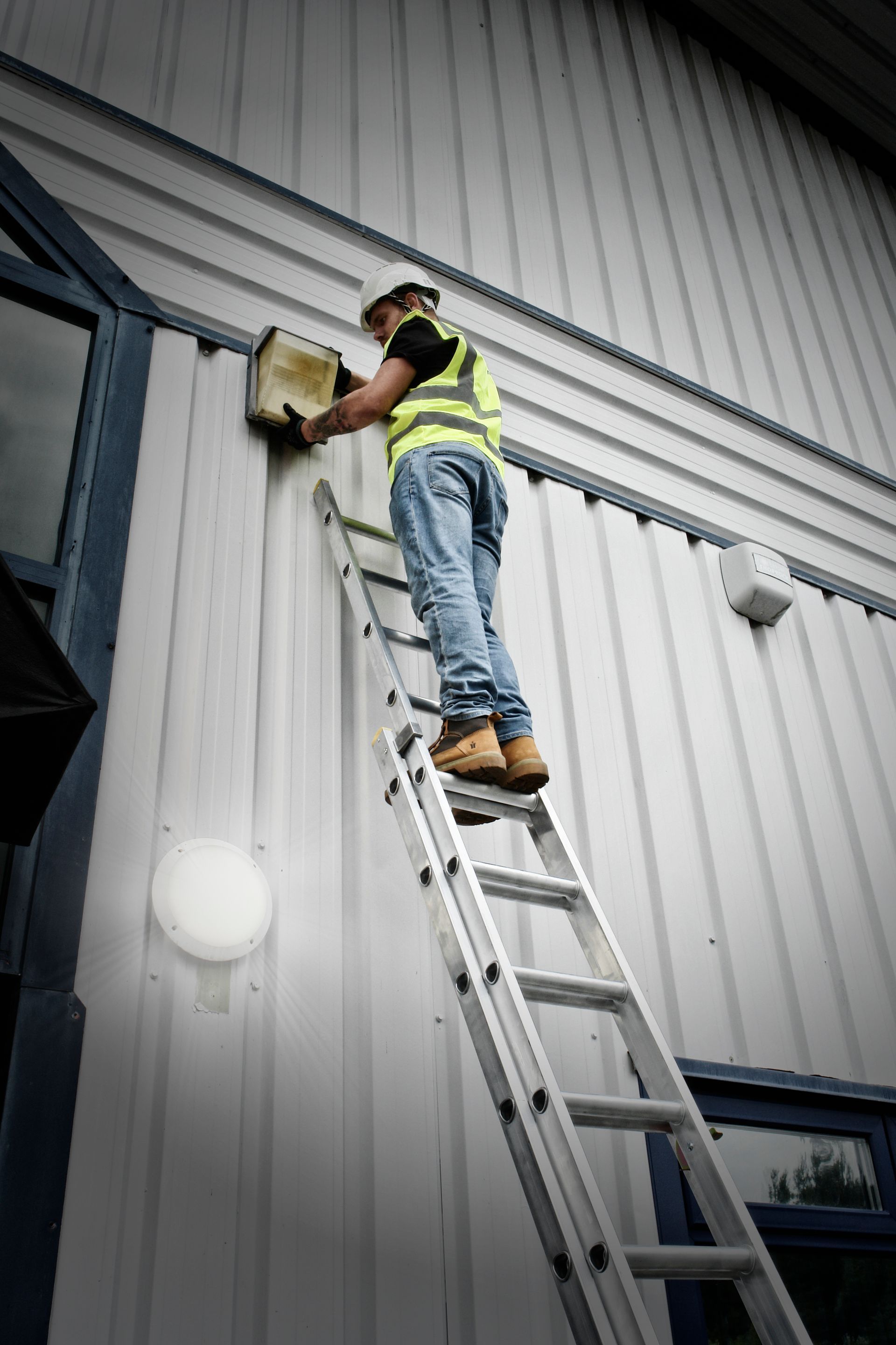 Single worker maintaining an external light fitting on a corrugated industrial building
