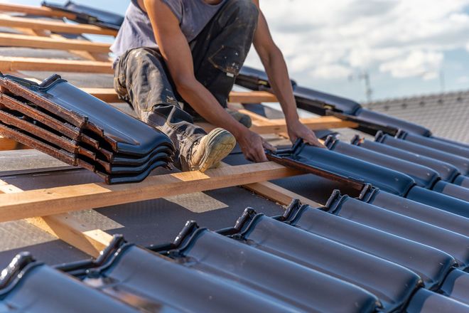 Roofer installing dark ceramic tiles on a wooden roof frame.