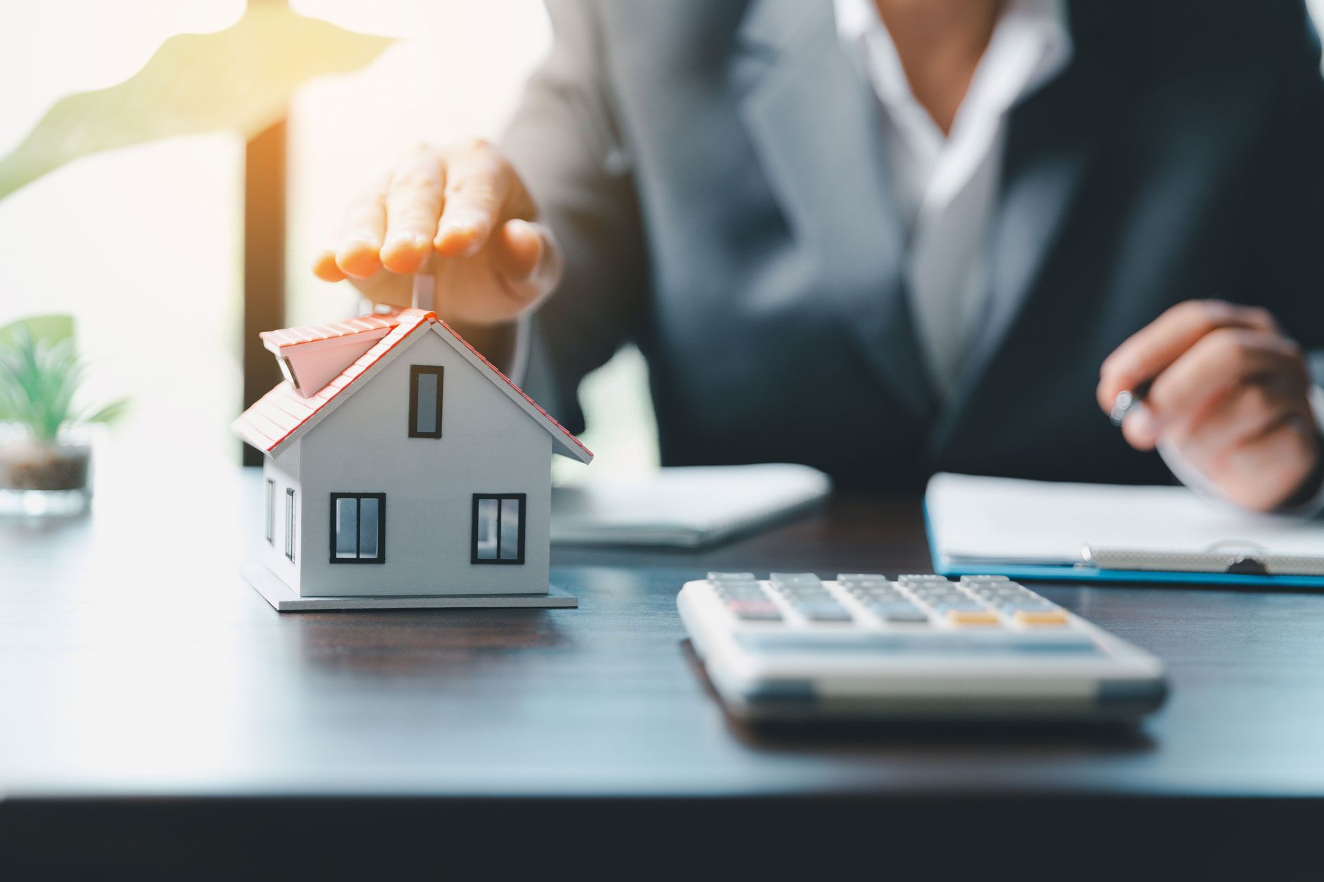 Person in suit protecting a model house with their hand, next to calculator and paperwork.