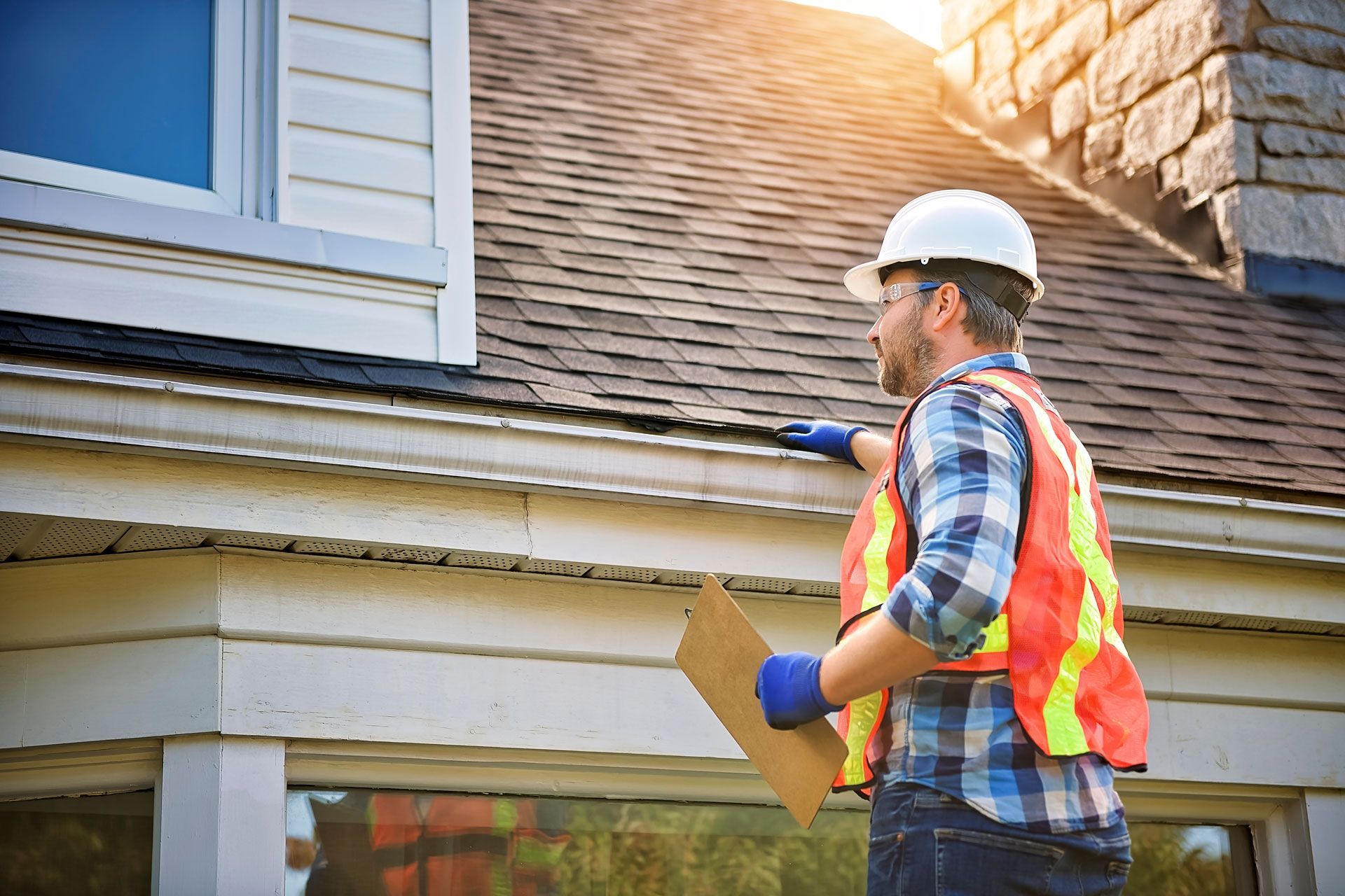 Construction worker in hard hat inspecting a gutter on a house, pointing with clipboard in hand.