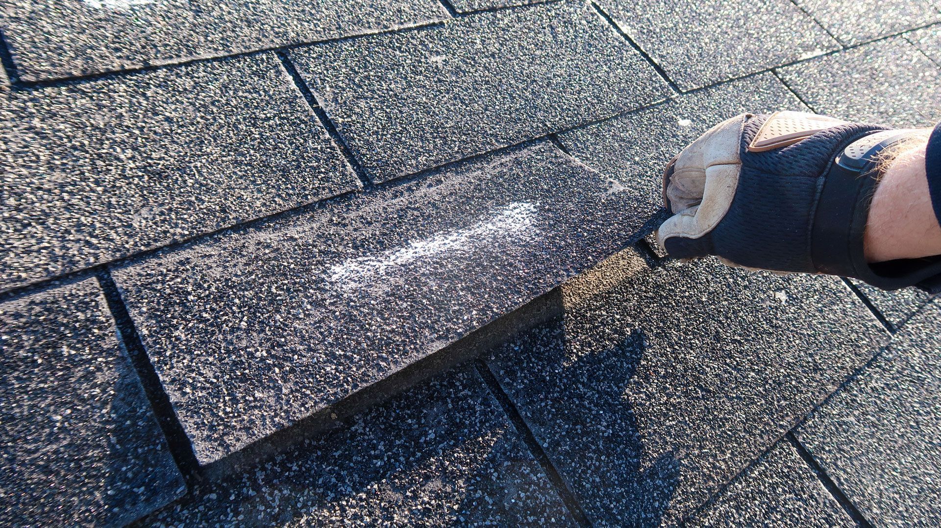 Hand with a glove holding up a gray shingle on a textured asphalt roof.