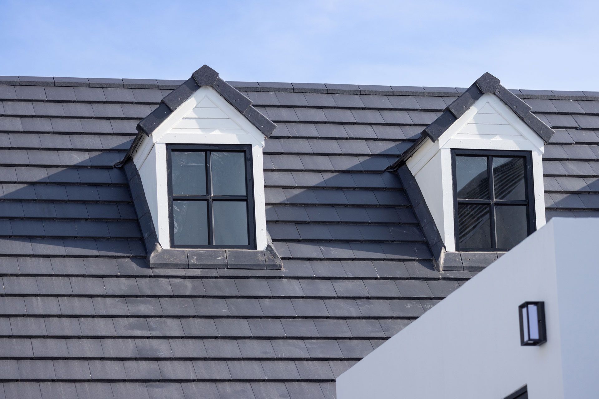 Two white dormer windows with black-framed windows on a gray tiled roof; partial white building.