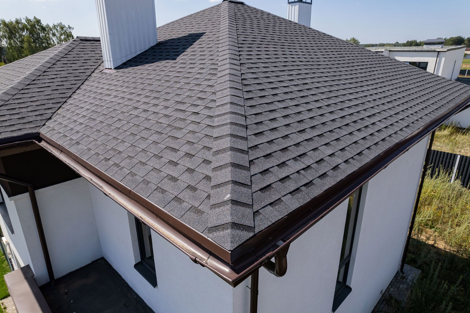 Dark gray shingle roof on a white house, brown gutters, chimneys, and surrounding grass.
