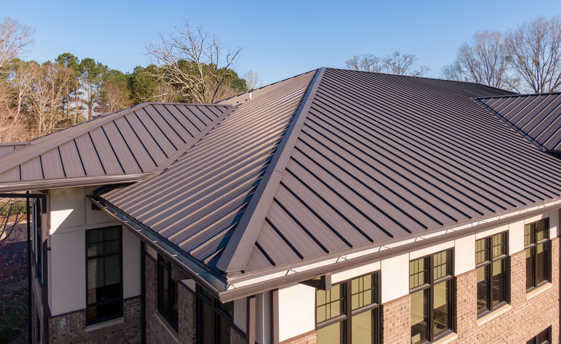 Dark metal roof on a building with brick and white accents, surrounded by trees under a blue sky.