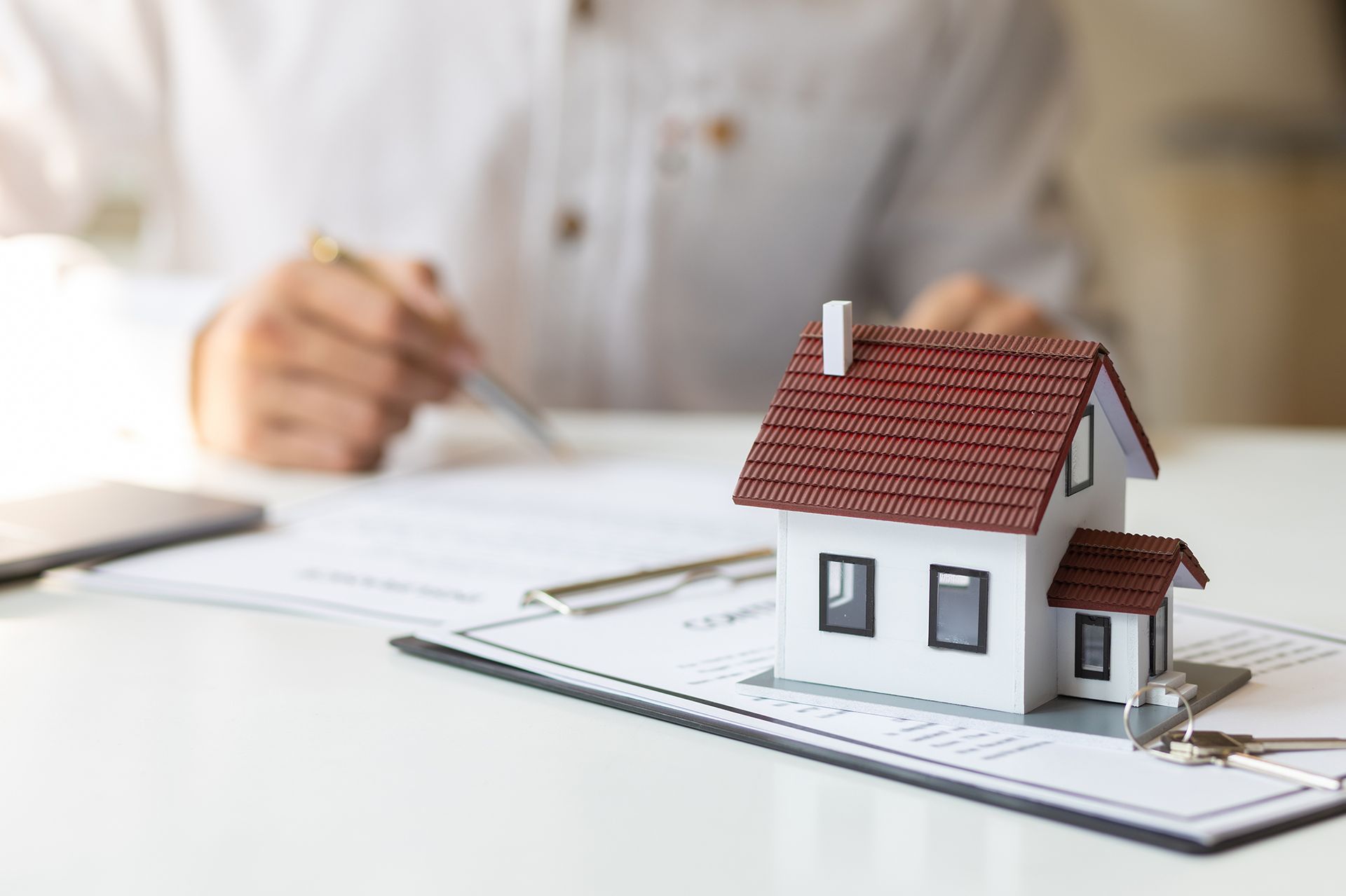 Miniature house on a clipboard with documents, person in white shirt writing in background.