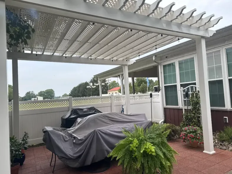 White pergola over patio with covered grill and table; ferns and potted plants.