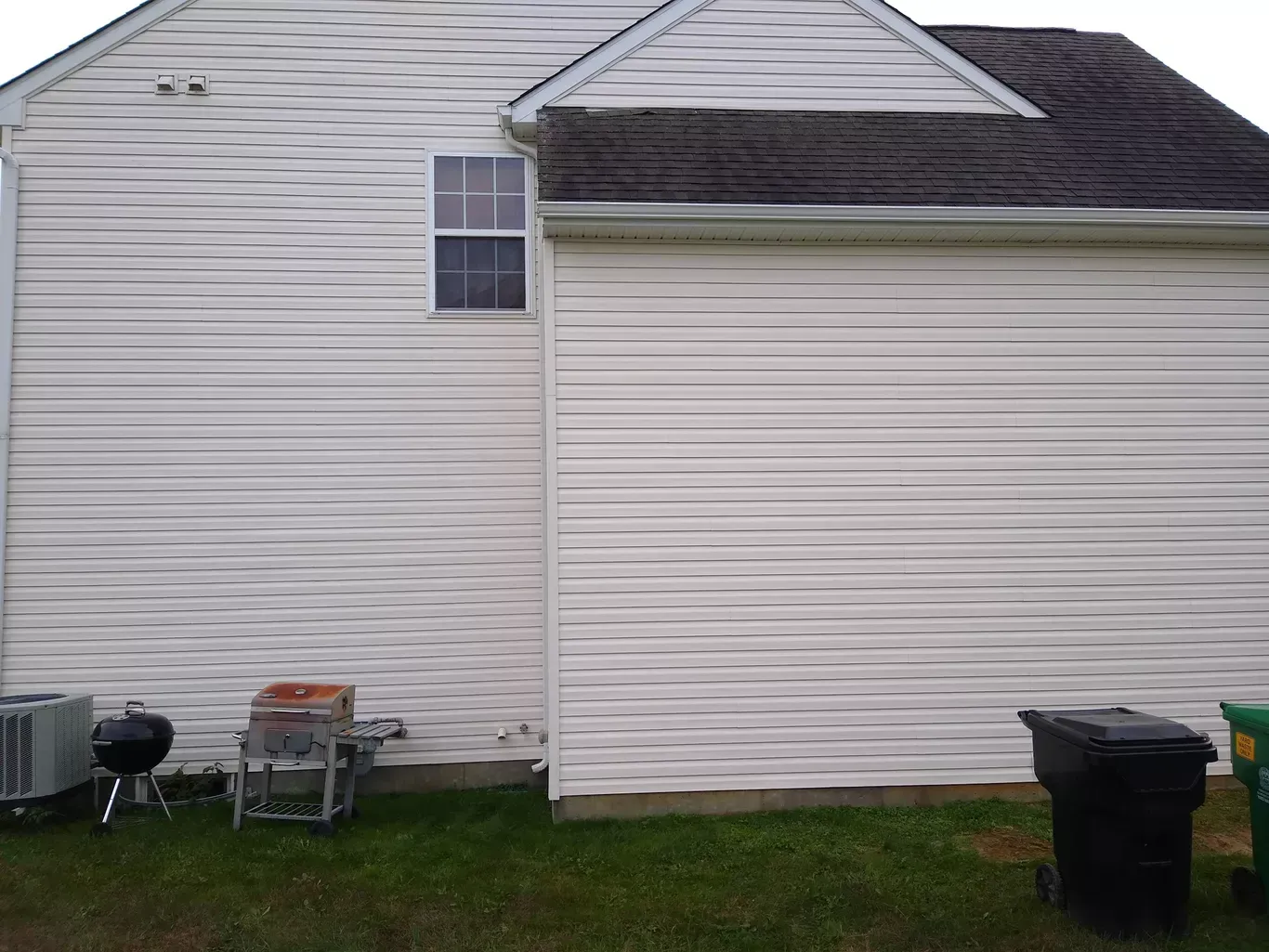 Side view of a house with white siding, a window, and a black trash bin on the grass.