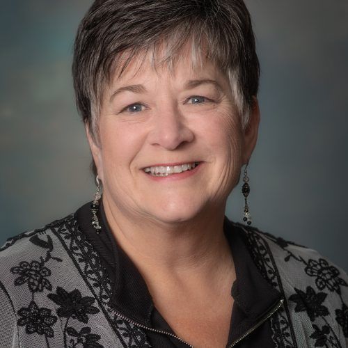 A woman wearing a black shirt and a gray jacket is smiling for the camera.