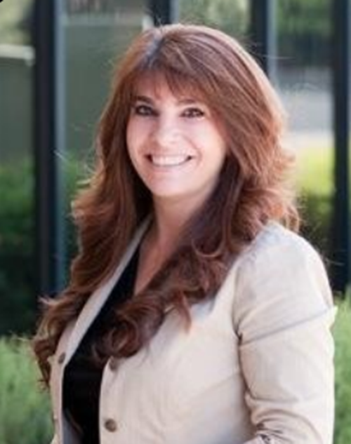 A woman with long red hair is wearing a tan jacket and smiling.