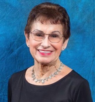 A woman wearing glasses and a necklace is smiling in front of a blue background.
