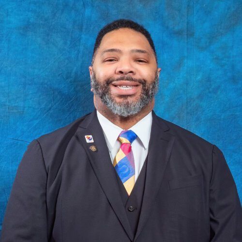 A man in a suit and tie is smiling in front of a blue background.