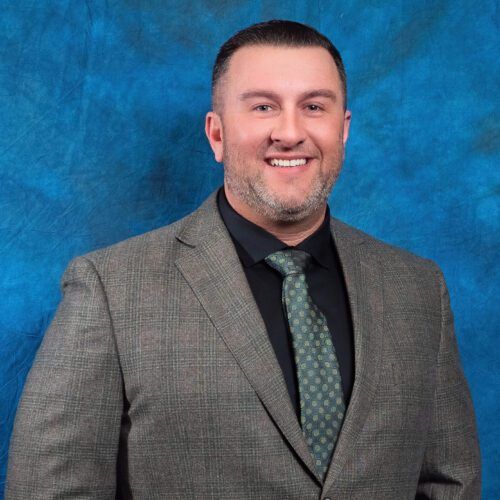 A man in a suit and tie is smiling in front of a blue background.