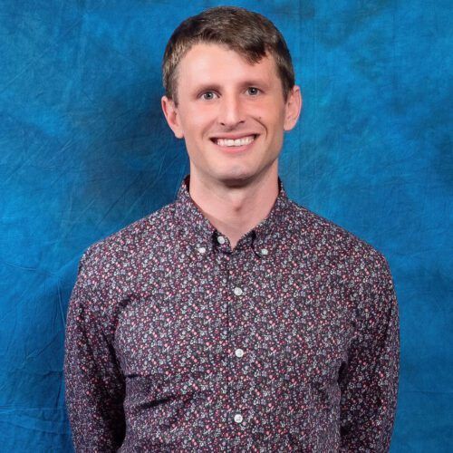 A man in a floral shirt is smiling in front of a blue background.