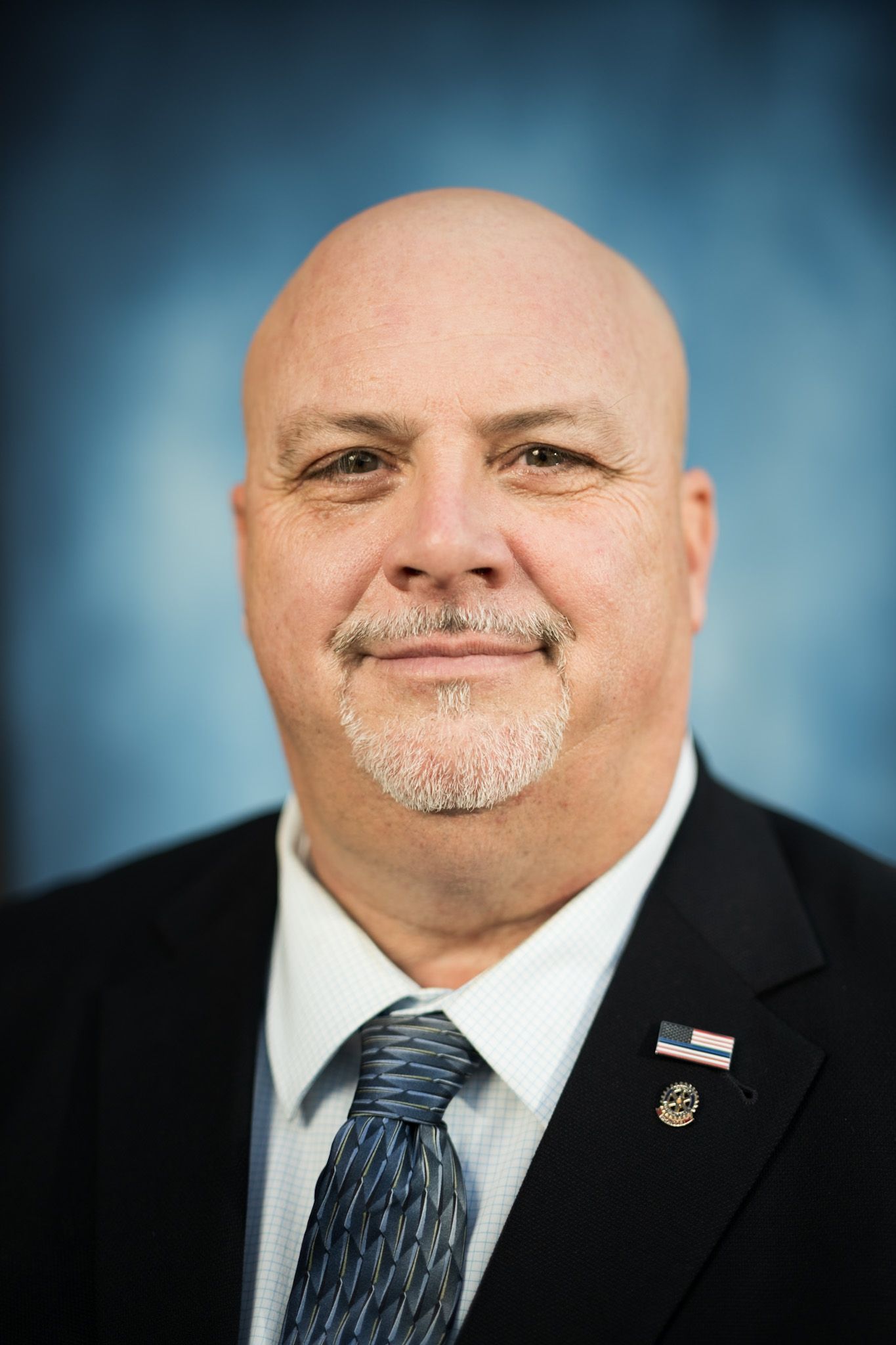 Bald man with goatee in suit, looking directly at the camera, against a blue background.