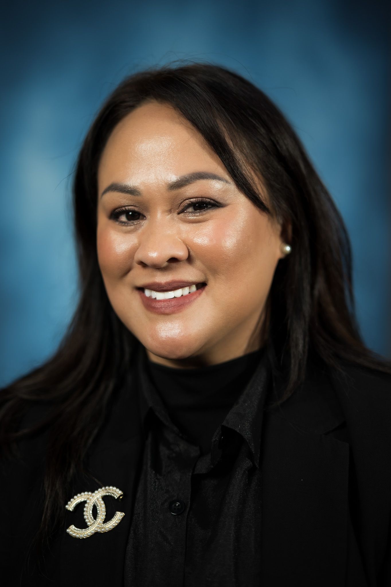 Woman with dark hair smiles, wearing a black blazer and turtleneck, with a crystal brooch.