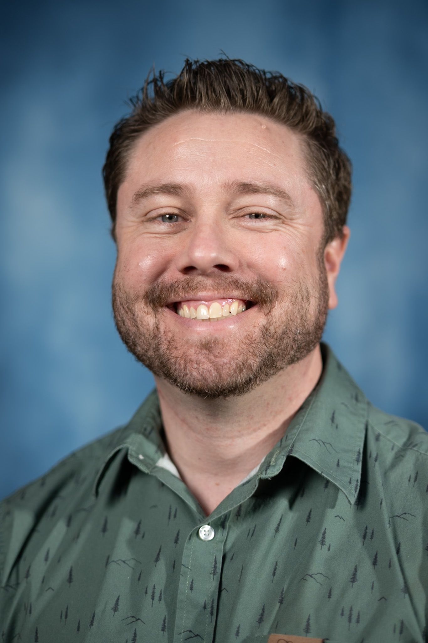 Man with a beard smiling, wearing a green patterned shirt against a blue background.