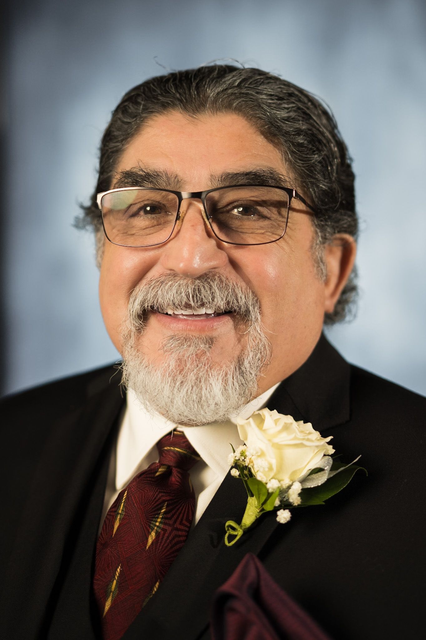 Man in suit with glasses, a white rose corsage, and a burgundy tie smiles.