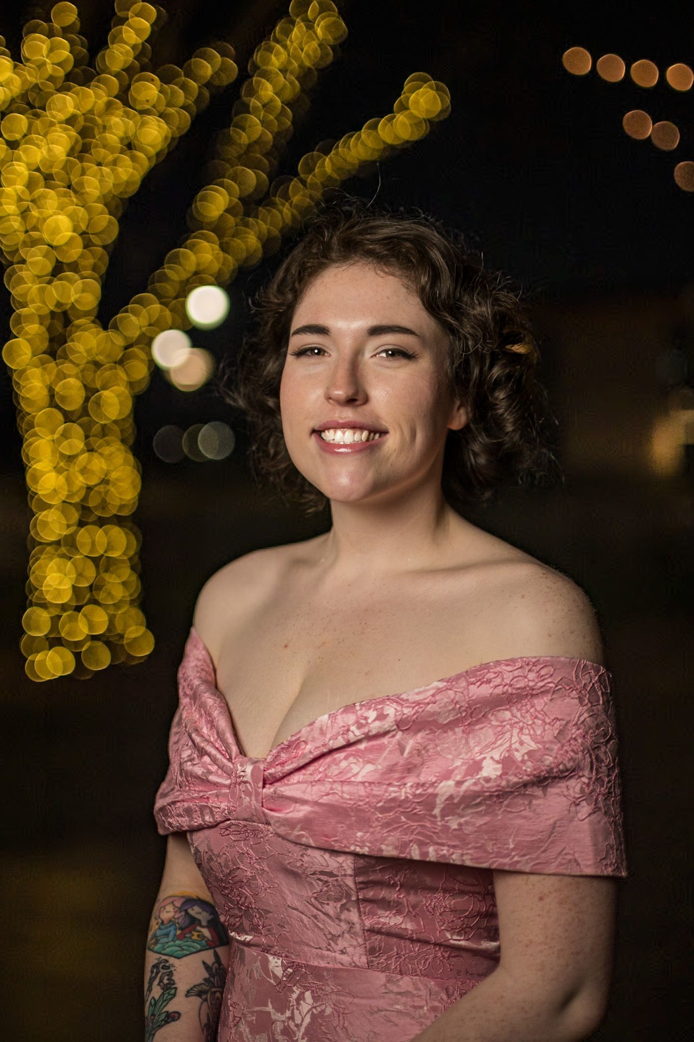 Woman in a pink off-the-shoulder dress smiles. Nighttime setting with blurred yellow lights in the background.