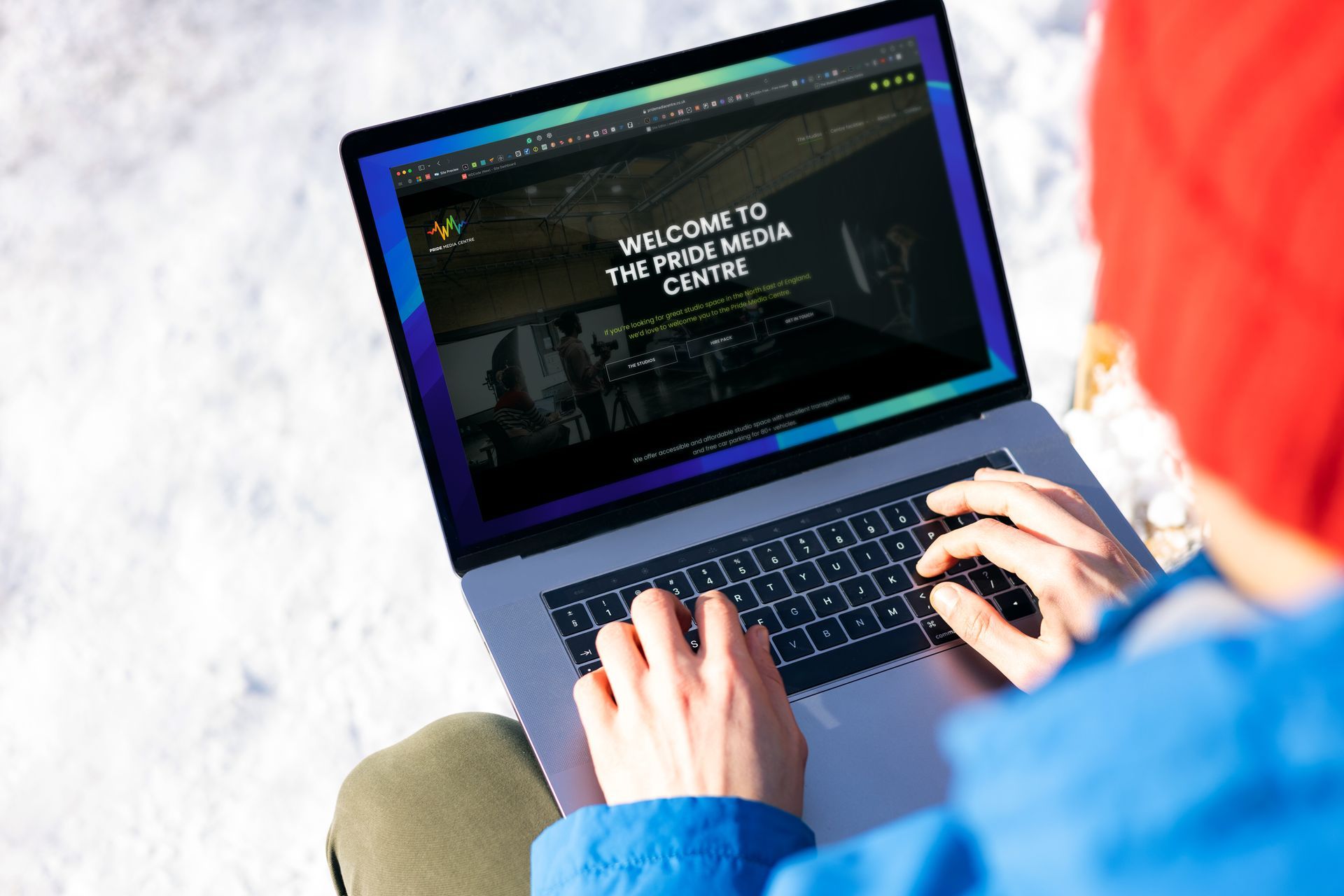 Person typing on a laptop outdoors in the snow, viewing a website. Red hat, blue jacket.