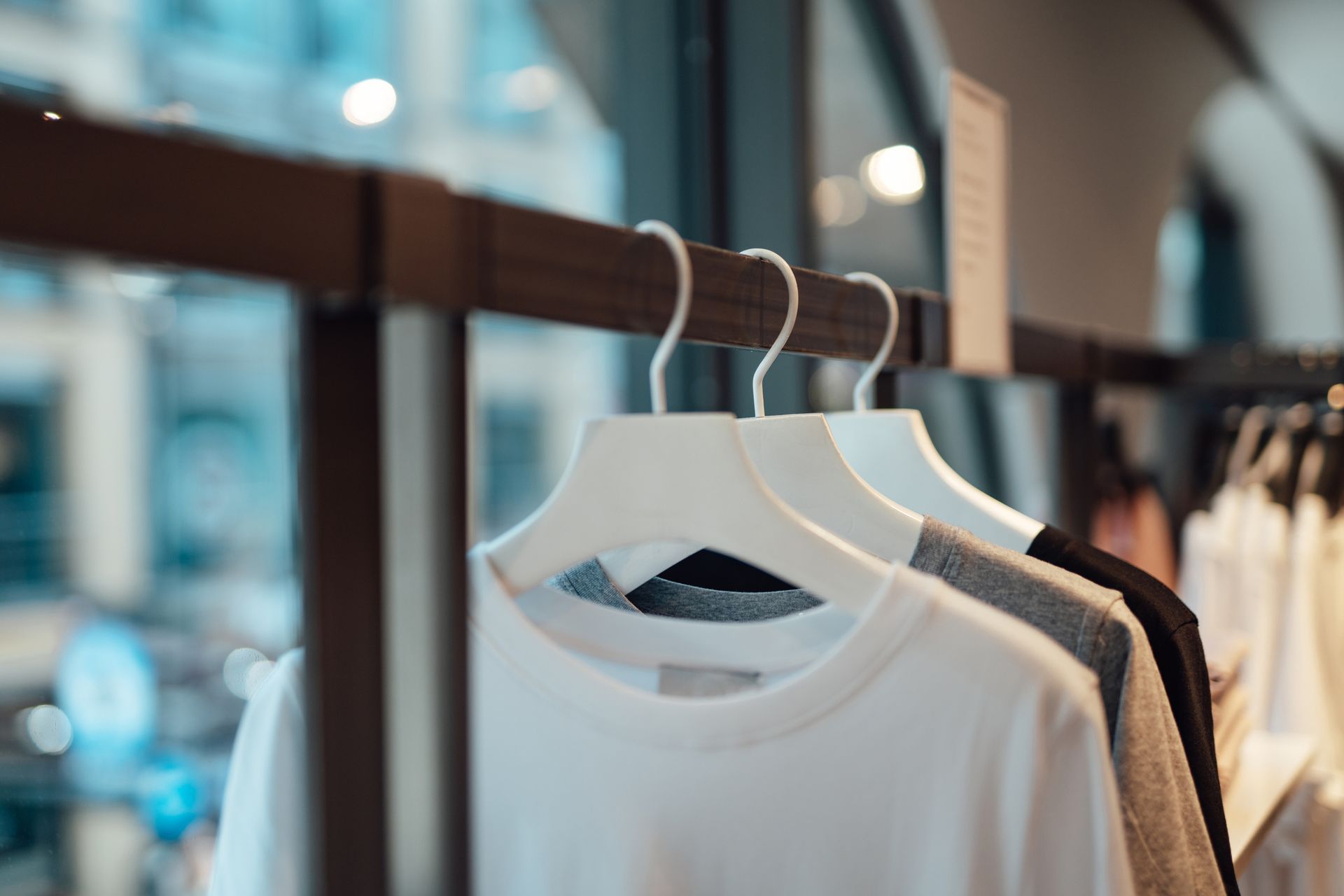 White, gray, and black shirts hanging on a rack inside a store, with blurred city view in background.