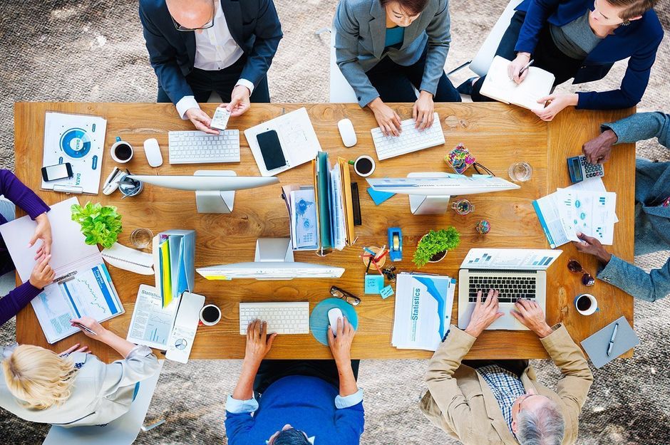 Group of people working collaboratively around a wooden table covered with papers, computers, and coffee cups in an office setting.