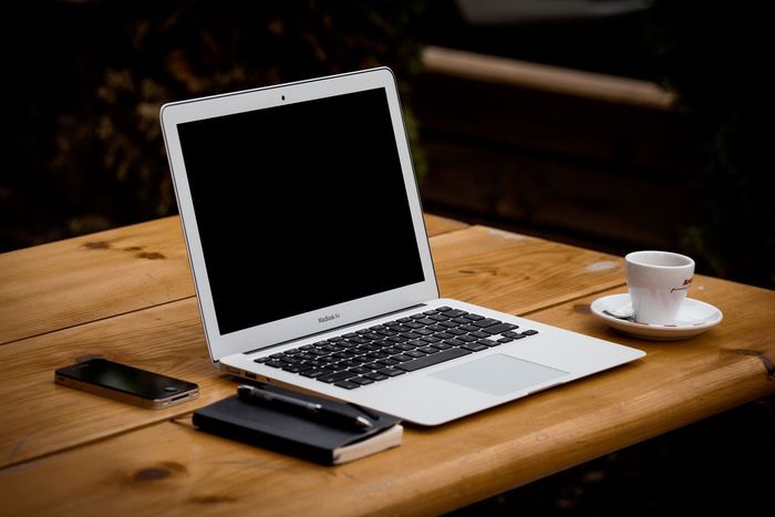 Laptop, smartphone, notebook, pen, and coffee cup on a wooden table outdoors. Screen is blank.
