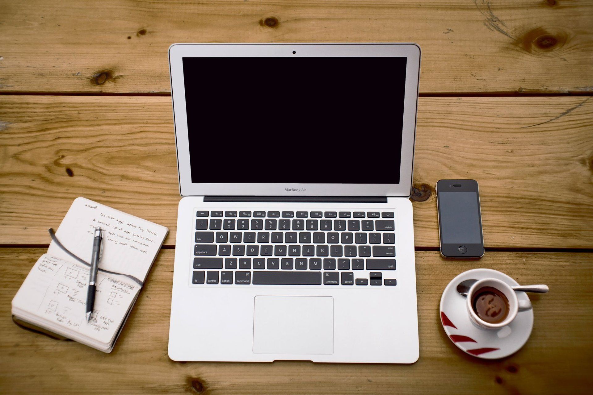 Laptop on table with a pen and paper on the left and a mobile phone and cup of coffee on the right