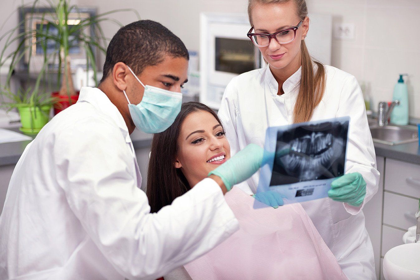 A woman is sitting in a dental chair while two dentists look at an x-ray of her teeth.