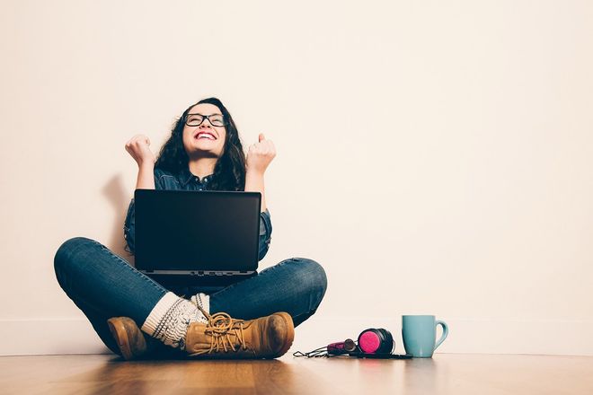 Woman with glasses, joyful expression, seated on floor with laptop, hands raised, coffee cup, headphones.