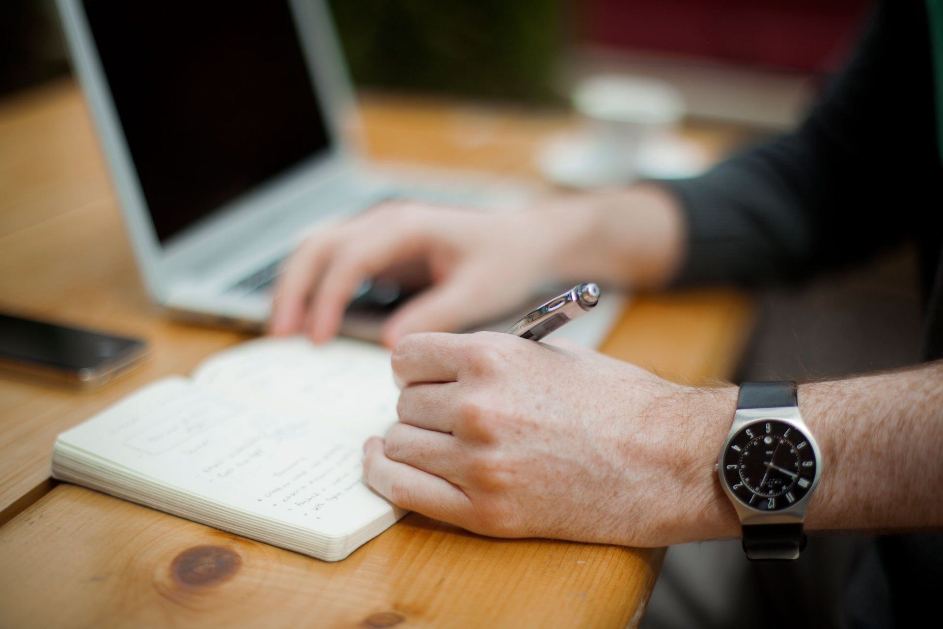 A man is writing in a notebook while using a laptop computer.