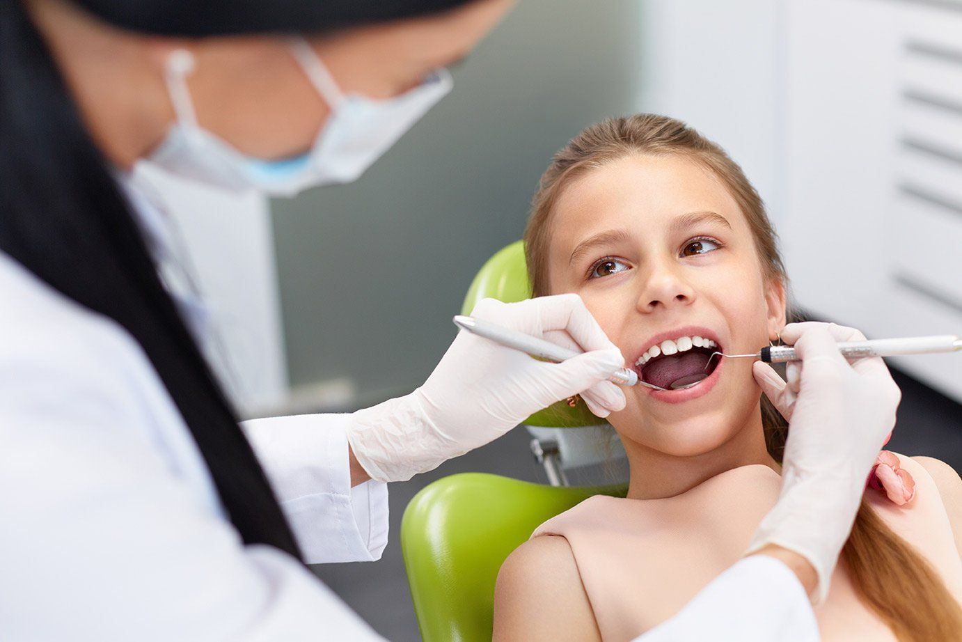 Preventative Dentistry for the Whole Family - a young girl is sitting in a dental chair getting her teeth examined by a dentist .