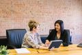 Two women at a table looking at a tablet, talking. Brick wall background.
